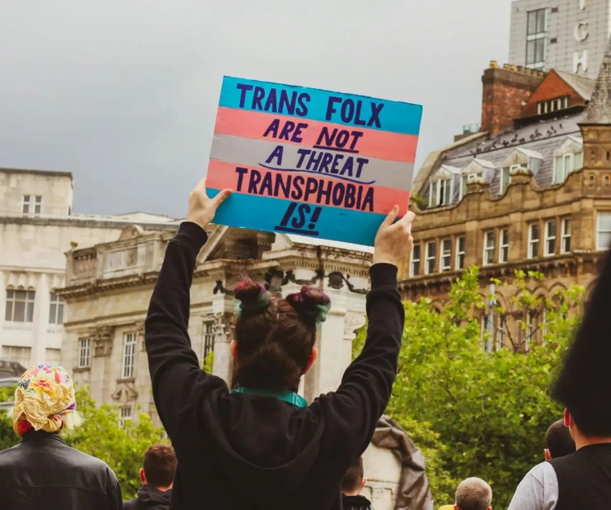 Tijdens een mars houdt iemand een bord omhoog met de Engelse tekst “trans mensen zijn geen bedreiging, transfobie is dat wel”.