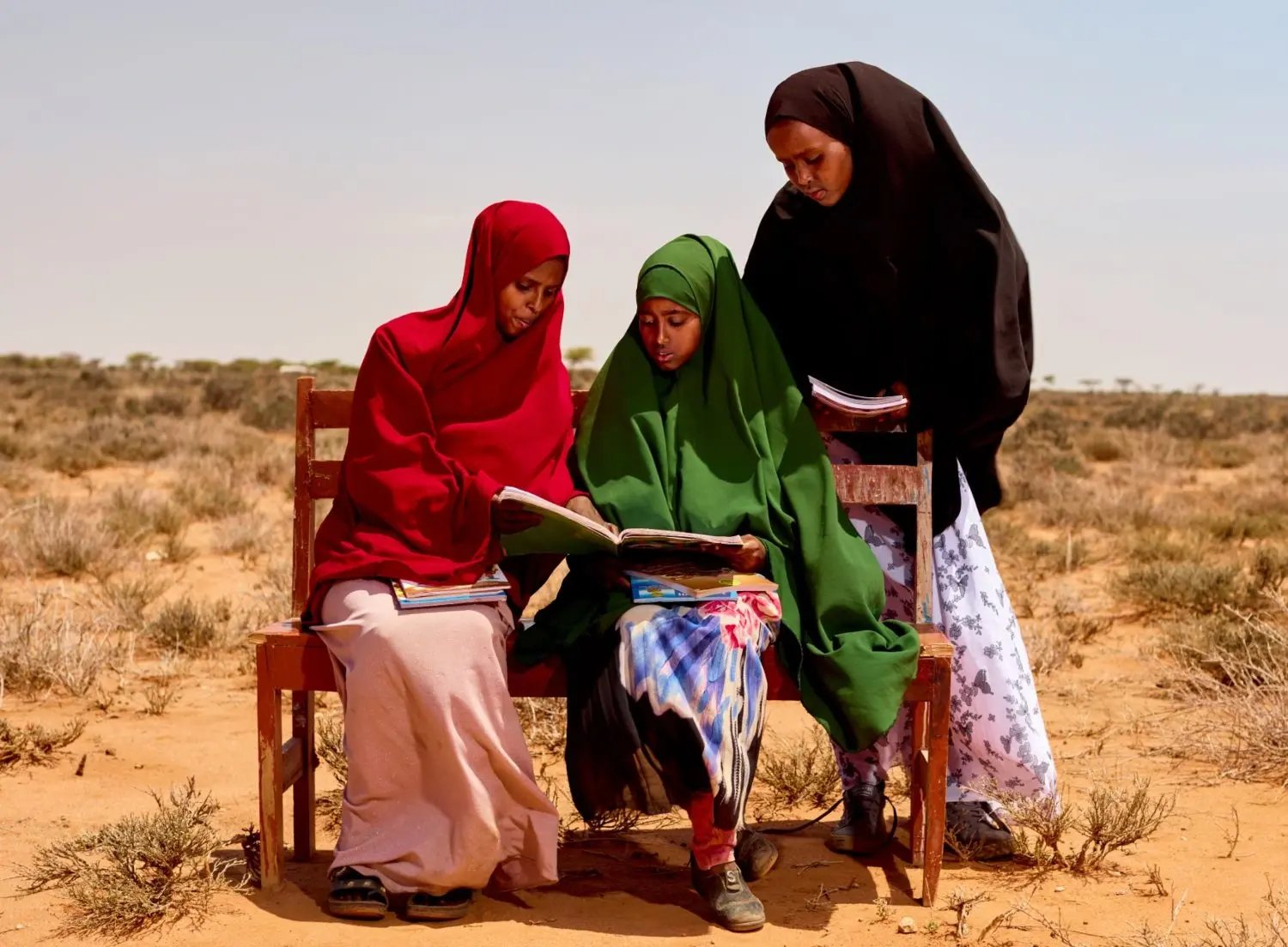 Hibbaq, ZamZam et Juweriya lisent un livre sur un banc dans le désert en Somalie. @Plan International/Armstrong Too