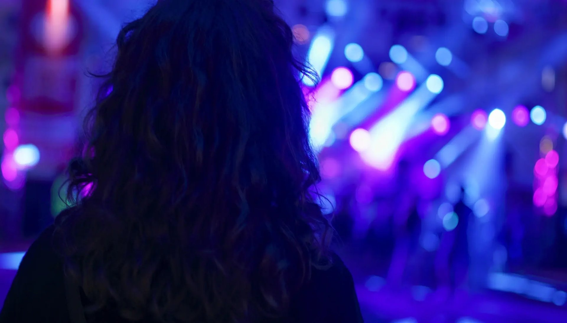 Une fille de dos, dans l’obscurité, arrive dans une fête nocturne animée avec des lumières. 