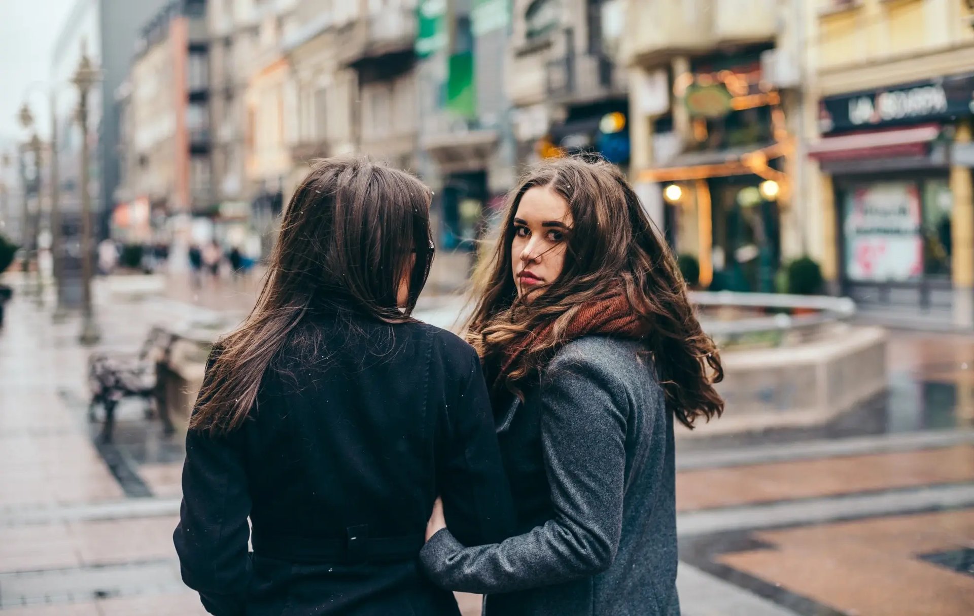 Twee jonge vrouwen wandelen in een stadscentrum, arm in arm. Een van hen draait zich om. (Credits: iStock)