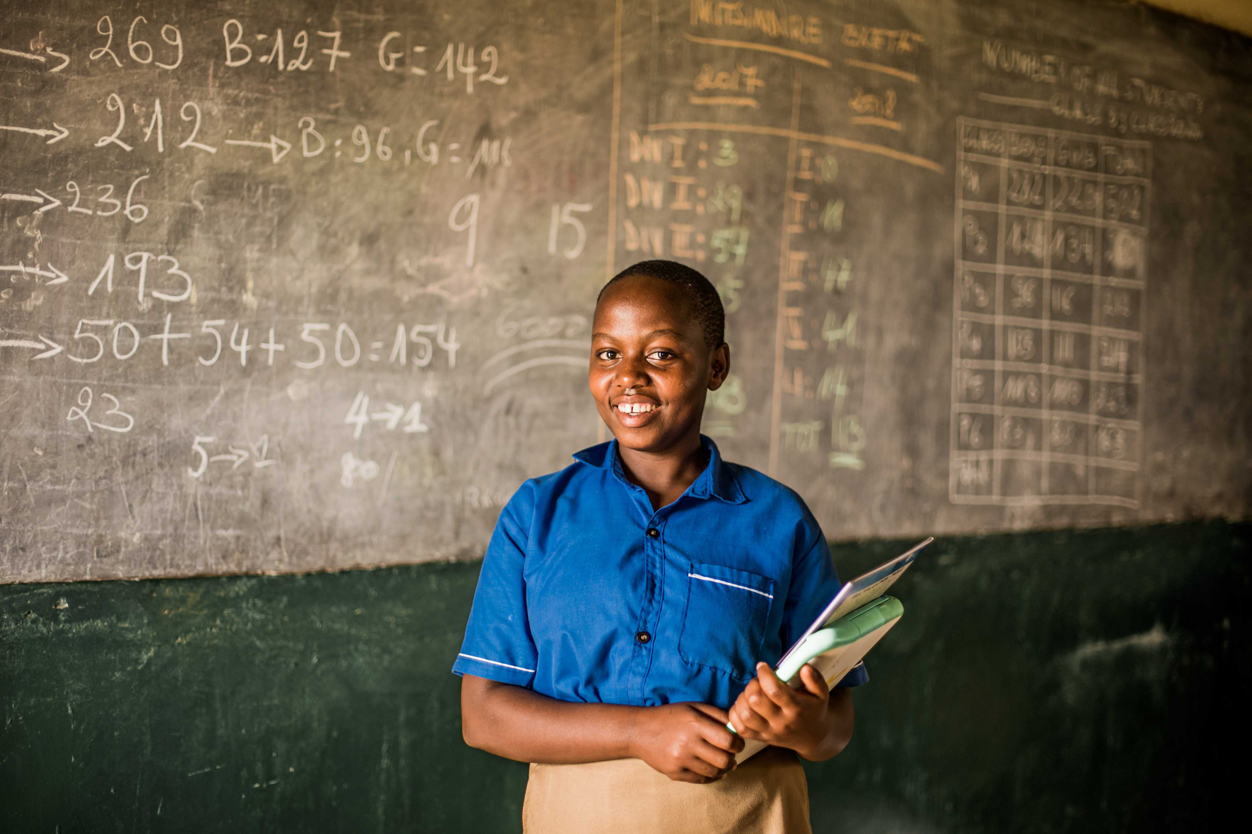 Ecolière au Rwanda sourit à la caméra devant le tableau de classe, un cahier à la main.